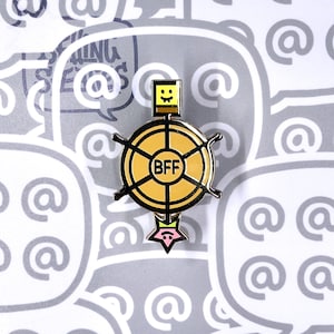 Puede incluir: Un pin de esmalte amarillo y negro con un diseño de robot de dibujos animados. El pin presenta una cabeza cuadrada con una cara sonriente, un cuerpo circular con "BFF" en el centro y una base en forma de estrella. El fondo es un patrón de burbujas de diálogo y símbolos @.