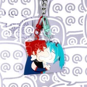May include: A double-sided acrylic keyring featuring two stylised anime characters with red and teal hair, connected by a silver metal chain with red and mint green lobster clasps. The characters are depicted in a close embrace, with visible details like dark blue clothing and white accents on a clear acrylic base.