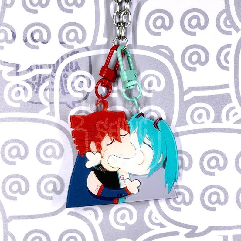 May include: A double-sided acrylic keyring featuring two stylised anime characters with red and teal hair, connected by a silver metal chain with red and mint green lobster clasps. The characters are depicted in a close embrace, with visible details like dark blue clothing and white accents on a clear acrylic base.