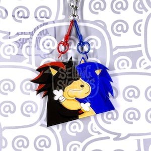 May include: A double-sided acrylic keychain featuring two characters, one black with red accents and one blue, facing each other. They are connected by a silver metal ring with a red lobster clasp on the left and a blue lobster clasp on the right.