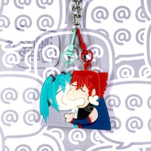 May include: A double-sided acrylic keyring featuring two cartoon characters kissing. One character has teal hair and the other has red hair. They are connected by a silver metal chain with a light blue clasp on the left and a red clasp on the right.