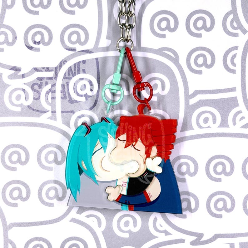May include: A double-sided acrylic keyring featuring two cartoon characters kissing. One character has teal hair and the other has red hair. They are connected by a silver metal chain with a light blue clasp on the left and a red clasp on the right.