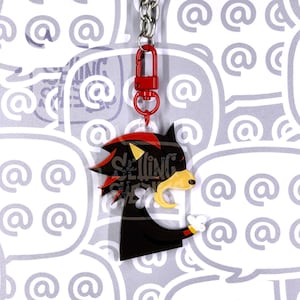 May include: An acrylic keychain featuring a stylized black character with red highlights, a yellow muzzle, and a white gloved hand. The character is attached to a red lobster clasp and a silver metal chain. The design includes a watermark overlay.