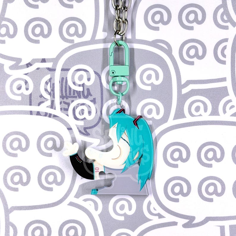 May include: An acrylic keyring featuring a stylised cartoon character with teal hair in twin tails, wearing a black and white outfit with the number 01. The charm is attached to a light green lobster clasp and a silver metal chain link.