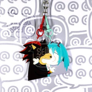 May include: A double-sided acrylic keyring featuring two characters joined together. One side shows a black and red hedgehog character, while the other side shows a teal-haired character. The keyring includes a silver chain and two clasps.