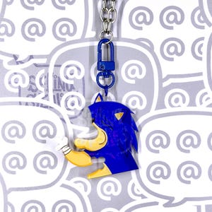 May include: A blue and yellow acrylic keychain featuring a stylized character head with spiky hair, a tan muzzle, and a white gloved hand. The charm is attached to a blue plastic lobster clasp and a silver metal chain, hanging vertically.
