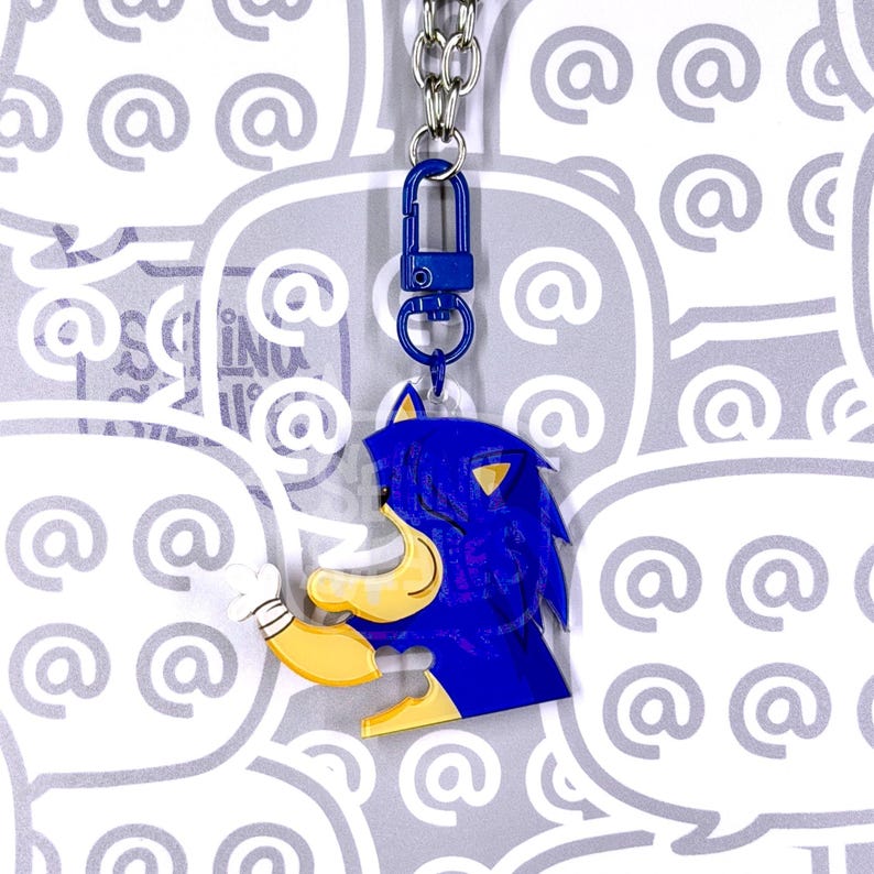 May include: A blue and yellow acrylic keychain featuring a stylized character head with spiky hair, a tan muzzle, and a white gloved hand. The charm is attached to a blue plastic lobster clasp and a silver metal chain, hanging vertically.