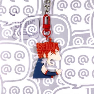 May include: An acrylic keyring featuring a stylised character with bright red hair and a dark blue outfit. The character's hair contains the word SELLING in bold letters. The keyring is attached to a red lobster clasp and a silver metal chain.