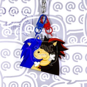 May include: A double-sided acrylic keychain featuring two interlocking character charms. One charm is blue with spiky hair, and the other is black with red highlights and white gloves. They are connected by a silver chain with blue and red clasps.