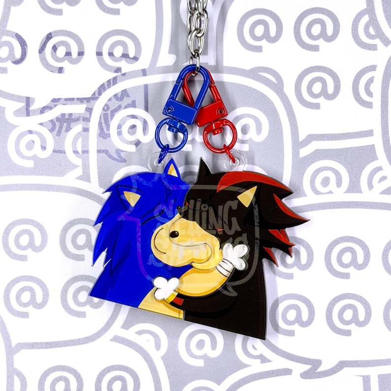 May include: A double-sided acrylic keychain featuring two interlocking character charms. One charm is blue with spiky hair, and the other is black with red highlights and white gloves. They are connected by a silver chain with blue and red clasps.