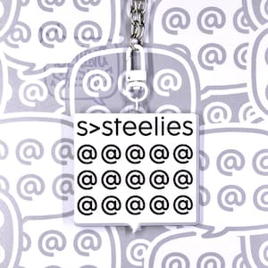 May include: A square white acrylic keychain featuring the black text s>steelies at the top, followed by a grid of fifteen black at symbols. The keychain is attached to a silver metal chain and a white plastic lobster clasp connector.