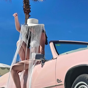 May include: A woman wearing a white fringe hat sits on the hood of a pink convertible car. The car is parked in front of a palm tree against a bright blue sky.