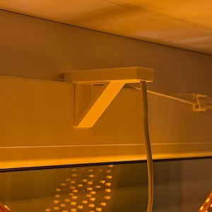 Puede incluir: Un soporte blanco en ángulo recto está montado en un marco de ventana blanco. Un cable blanco cuelga del soporte. La imagen está iluminada con una luz cálida y amarilla.