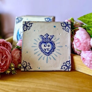 Può includere: Una piastrella di ceramica bianca con un design blu e bianco di un cuore con una corona e la parola "LOVE" all'interno. La piastrella ha un bordo blu e bianco con un motivo floreale.