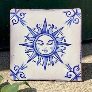 Peut inclure: Carreau de céramique carré avec un motif de soleil peint à la main en bleu sur fond blanc. Le soleil a les yeux fermés et un visage souriant. Des volutes bleues décoratives sont dans chaque coin.