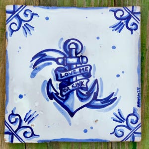 Könnte beinhalten: Quadratische Keramikfliese mit handgemaltem blauem Ankerdesign und den Worten "LOVE ME OR SINK" auf einem Banner. Die Fliese hat einen weißen Hintergrund mit blauen dekorativen Eckakzenten und einem blauen Rand. Die Fliese liegt auf einer grünen Holzoberfläche.