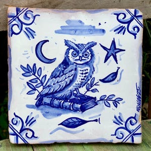 May include: Square ceramic tile featuring a hand-painted owl design in blue hues. The owl sits on books, with a crescent moon, star, and leaves. Decorative blue border and corner accents.