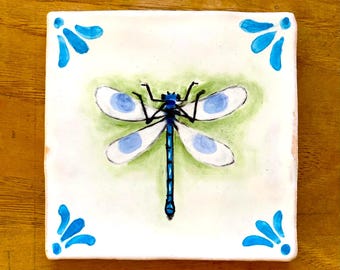 May include: A square ceramic tile featuring a hand-painted blue dragonfly centered on a soft green wash background. The four corners are decorated with blue floral-inspired motifs. The tile has a white base and a smooth, glossy finish.