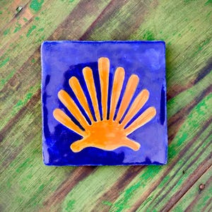 May include: Square blue ceramic tile with an orange scallop shell design. The tile is set against a green and brown wooden surface. The shell design is a symbol of the Camino de Santiago pilgrimage.