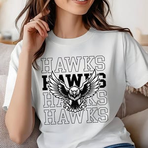以下が含まれることがあります： 「HAWKS」の文字が縦に繰り返され、中央に白黒のワシのグラフィックが描かれた白いTシャツ。ワシは翼を広げています。Tシャツは柔らかい素材で作られています。デザインはTシャツの中央に配置されています。