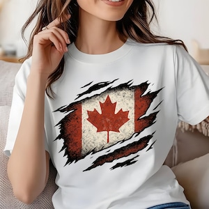 May include: White t-shirt featuring a distressed Canadian flag design. The flag includes a red maple leaf on a white background, framed by red and black tear marks. The shirt is a casual wear item.