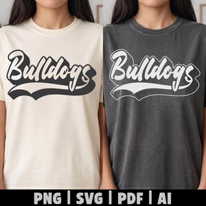 May include: Two short-sleeve crew neck t-shirts, one in cream with a dark grey graphic and one in charcoal grey with a white graphic. Both shirts feature the word Bulldogs in a stylised, bold, cursive script font with a drop shadow effect.