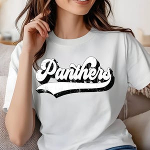 Peut inclure: T-shirt blanc avec le mot "Panthers" dans une police rétro et vieillie. Le texte est blanc avec un contour noir et une bannière noire en dessous. Le t-shirt est de style col rond classique.