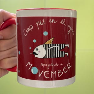 Może przedstawiać: Czerwono-biały ceramiczny kubek z czerwonym uchwytem. Kubek przedstawia kreskówkową rybę z wąsami oraz napisy "Como pez en el agua" i "Apoyando a NOVEMBER" w białym skrypcie. Niebieskie akcenty w postaci bąbelków.