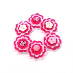 May include: A collection of felt flower embellishments in shades of pink and white, arranged in a circular pattern. Some flowers feature button or ladybug center details. The flowers are layered, creating a three-dimensional effect.