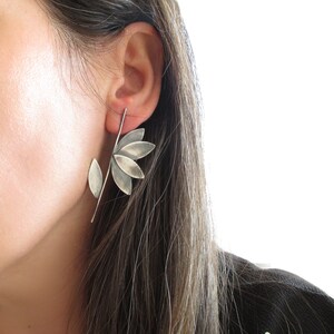 May include: A silver flower-shaped earring with a stem and a single leaf. The earring is worn in the left ear of a person with long brown hair.