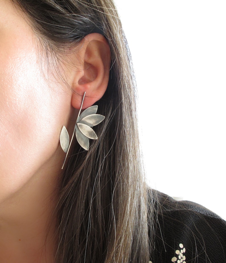 May include: A silver flower-shaped earring with a stem and a single leaf. The earring is worn in the left ear of a person with long brown hair.