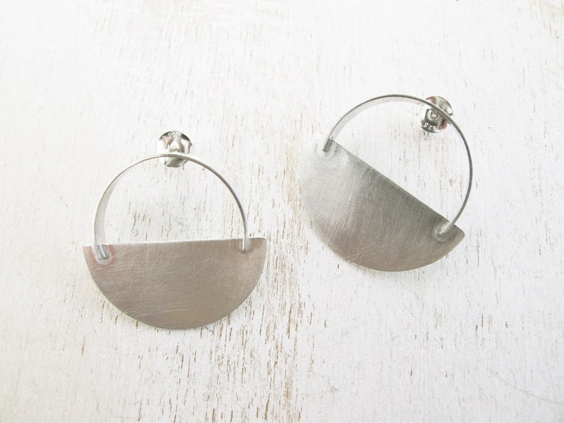 May include: A pair of silver earrings with a half-circle design. The earrings are made of a brushed metal and have a simple, modern style.
