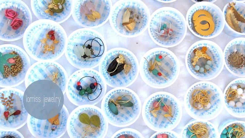 May include: Assortment of earrings displayed in white cupcake liners on a white background. The earrings are made of various materials, including metal, beads, and feathers. The text "iomiss jewelry" is visible in the center of the image.