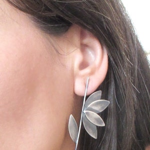 May include: Silver earrings with a leaf design. The earrings are long and dangle from the earlobe. The leaves are made of metal and have a textured surface.