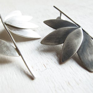 May include: A pair of silver earrings with a leaf design. The earrings are made of metal and have a brushed finish.