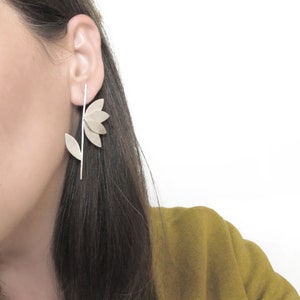 May include: Silver flower earrings with a long stem and multiple petals. The earrings are worn by a person with long dark hair.