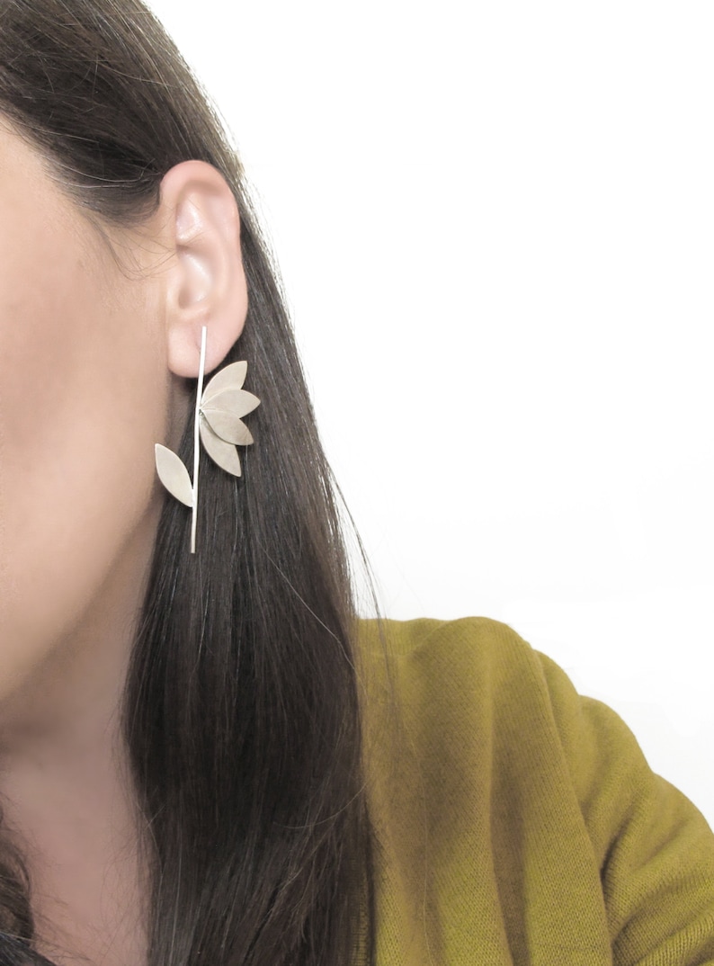 May include: Silver flower earrings with a long stem and multiple petals. The earrings are worn by a person with long dark hair.