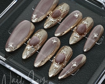 May include: Set of ten false nails with a light brown base and gold accents. Each nail has a large, oval-shaped, light brown gemstone with rhinestones around it.