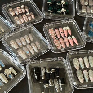 May include: A collection of ten clear plastic boxes containing various sets of artificial fingernails. Each set features different designs and colors, including pink, white, black, blue, and green. Some sets have rhinestones and other embellishments.