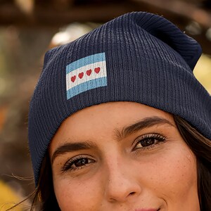 Puede incluir: Un gorro azul marino con un parche blanco y azul claro con tres corazones rojos. El parche es una versión estilizada de la bandera de Chicago.