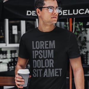 Puede incluir: Una camiseta negra con el texto "LOREM IPSUM DOLOR SIT AMET" impreso en gris en la parte delantera. La persona que lleva la camiseta sostiene una taza de café para llevar.
