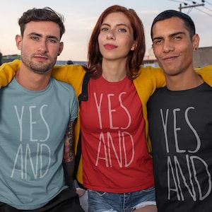 Puede incluir: Tres personas con camisetas a juego con el texto "YES AND" en blanco sobre camisetas de diferentes colores. La persona del medio lleva una camiseta roja, la persona de la izquierda lleva una camiseta azul claro y la persona de la derecha lleva una camiseta negra.
