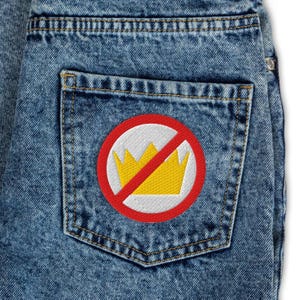 May include: Close-up of a denim jean pocket with a circular patch. The patch features a yellow crown inside a white circle, crossed out with a red diagonal line. The jeans have a light blue wash.