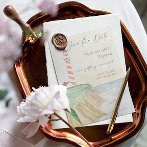Peut inclure: Carte de Save the Date avec une illustration aquarelle d'un phare et d'un paysage côtier. La carte est sur un plateau en cuivre avec un sceau de cire et un stylo. Le texte sur la carte est "Save the Date, Nolan et Trisha se marient, le 19 avril 2025, Chatham, MA."
