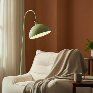 Może przedstawiać: Szałwiowa zielona lampa podłogowa z zakrzywionym ramieniem i kloszem w kształcie kopuły oświetla kremowy fotel. Beżowy koc jest zarzucony na krzesło, a zielony kubek stoi na małym drewnianym stoliku. Tło przedstawia brązową ścianę i okno.