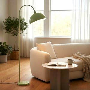 May include: A green arched floor lamp with a round base and a dome-shaped shade. The lamp is positioned next to a cream-colored sofa and a small round table. A book and a mug are on the table. The room has a bright, airy feel.