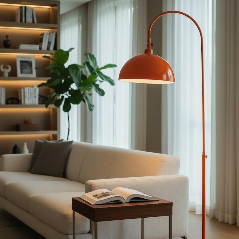 Może przedstawiać: Pomarańczowa lampa podłogowa z łukiem i kloszem w kształcie kopuły oświetla salon. Lampa stoi obok kremowej sofy i małego drewnianego stolika z otwartą książką. W tle regał i duża roślina.