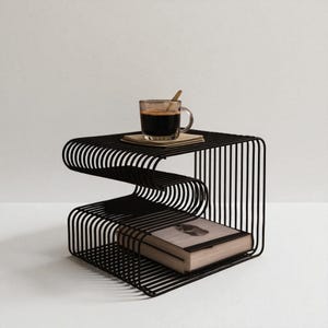 May include: A black metal side table with a unique, wavy design. A clear glass cup of coffee with a wooden spoon rests on top, along with a small stack of coasters. A book is placed on the lower shelf.