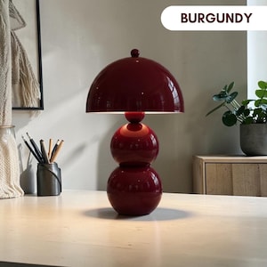 May include: A burgundy table lamp featuring a glossy finish, a dome-shaped shade, and a base composed of two stacked spheres. The lamp has a small knob on top of the shade. The text BURGUNDY is visible in the upper right corner of the image.