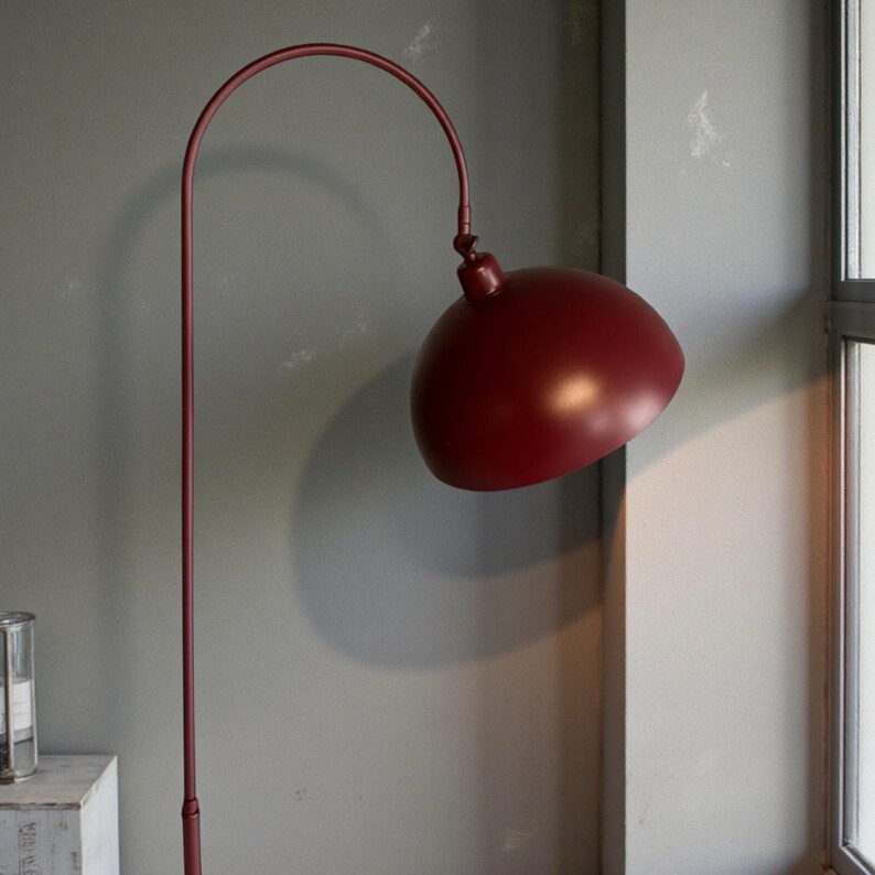 Może przedstawiać: Bordowa lampa podłogowa z zakrzywionym ramieniem i kloszem w kształcie kopuły. Lampa ma smukły, pionowy słupek i zaokrąglony klosz. Wysokość lampy to około 152 cm.